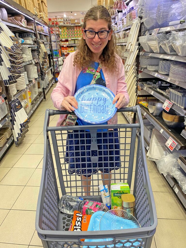 Person shopping for birthday party in a store aisle, holding a blue Happy Birthday plate above a trolley containing party supplies and groceries.